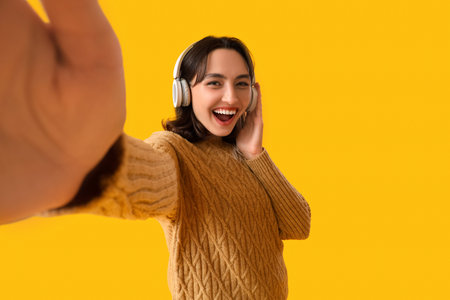 Young woman in warm sweater with headphones taking selfie on yellow background