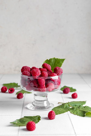 Glass bowl with fresh ripe raspberries and leaves on white tile table