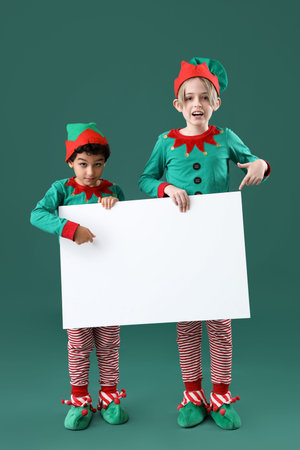 Cute little children in elves costumes with blank poster on green background