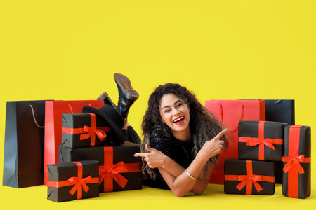Beautiful African-American woman with shopping bags and gift boxes on yellow background. Black...