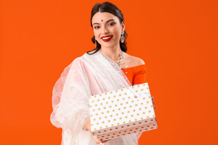 Beautiful young Indian woman in sari with gift box on orange background