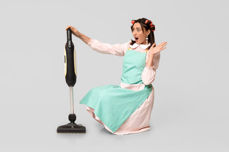 Portrait of shocked young housewife in apron with vacuum cleaner on gray backgroundの写真素材