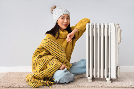 Young Asian woman in winter clothes with radiator near light wallの写真素材