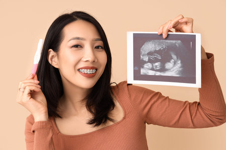 Young Asian woman with pregnancy test and sonogram image on beige background, closeup