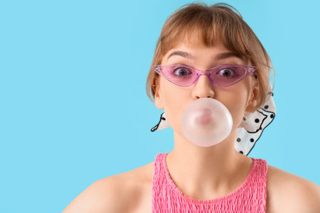 Beautiful young woman with chewing gum on blue background, closeup