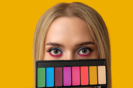 Beautiful young woman with palette of colorful eyeshadows on yellow background