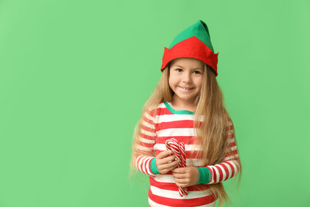 Cute little girl in elf costume with candy canes on green background