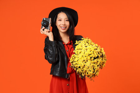 Young Asian woman with photo camera and chrysanthemum flowers on orange background