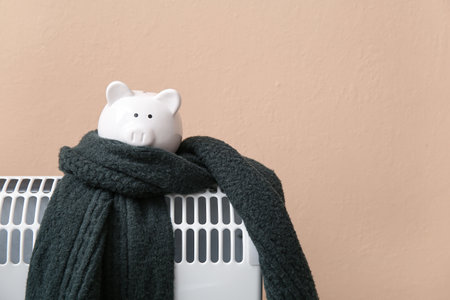 Piggy bank with warm scarf on electric convector heater near beige wall at home. Heating saving conceptの写真素材