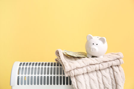 Piggy bank with warm blanket and money on electric convector heater near yellow wall at home. Heating saving conceptの写真素材