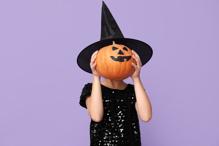 Cute little girl dressed for Halloween as witch with pumpkin on purple background
