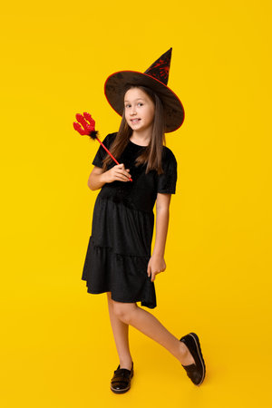 Cute little girl dressed for Halloween as witch with red devil pitchfork on yellow backgroundの写真素材