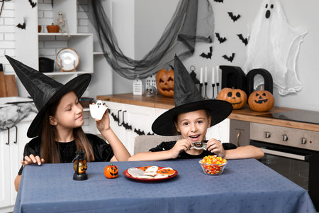 Cute little girls dressed for Halloween as witches with cookies and candies at table in kitchenの写真素材