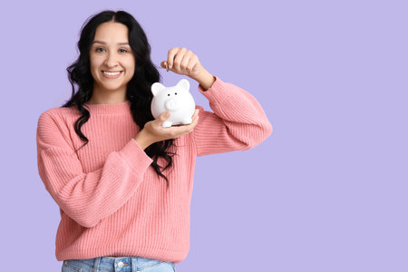 Pretty young woman putting coin into piggy bank on lilac backgroundの写真素材