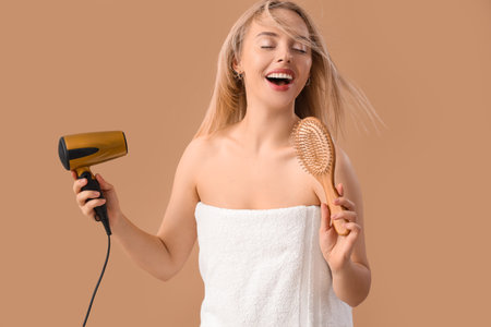 Young blonde woman with brush drying hair after shower on beige backgroundの写真素材