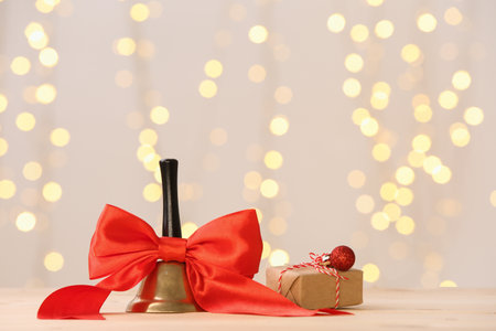 Christmas bell with red bow and gift box on white wooden table against blurred lightsの写真素材
