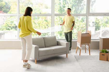 Happy young couple wrapping sofa in room on moving day
