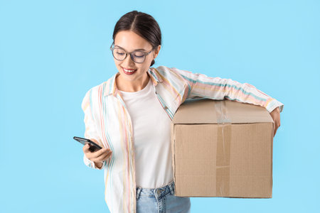 Young Asian woman with parcel using mobile phone on blue backgroundの写真素材