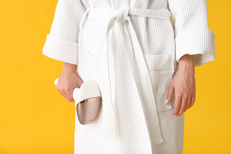 Beautiful young woman in bathrobe with photoepilator on yellow backgroundの写真素材