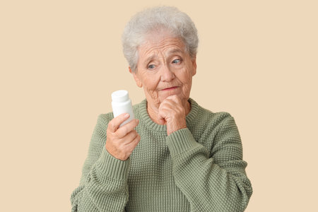 Thoughtful senior woman with pill bottle on beige backgroundの写真素材