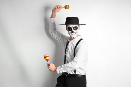 Young man with painted skull and maracas on light background. Mexico's Day of the Dead (El Dia de...