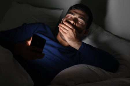 Sleepy young man using mobile phone in bed at night