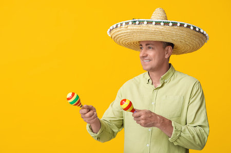 Handsome mature Mexican man in sombrero hat with maracas on yellow background