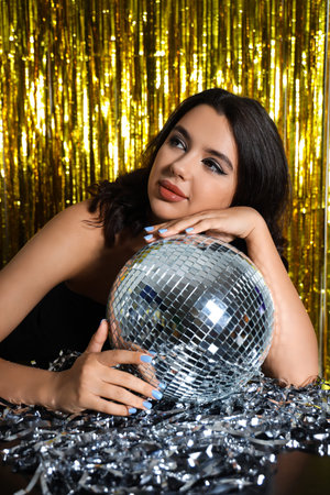 Beautiful young woman with disco ball against golden tinsel