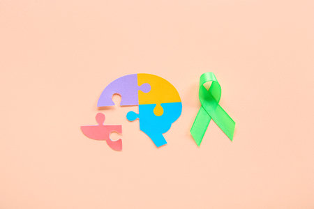 Colorful puzzle pieces in shape of brain with green awareness ribbon on pink background. Mental...