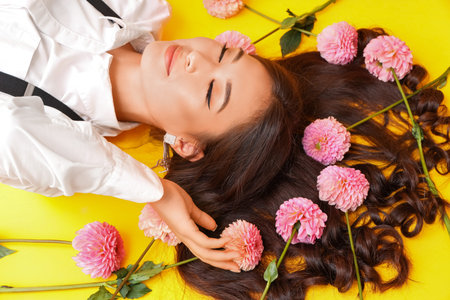 Beautiful young Asian woman with pink dahlia flowers in her hair on yellow background