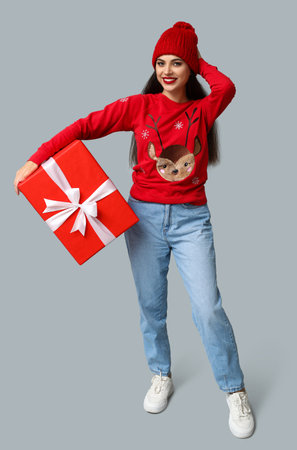 Beautiful young woman in Christmas sweater with gift box on gray backgroundの写真素材