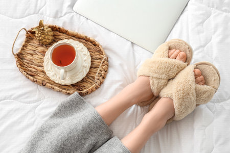 Young woman in soft slippers resting on bed with modern laptop and cup of teaの写真素材