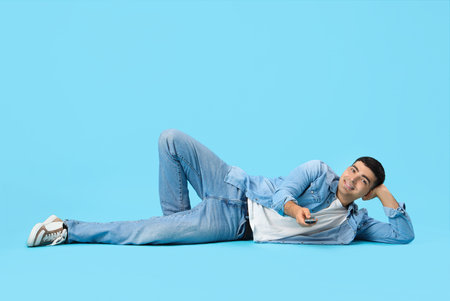 Handsome young man watching TV while lying on floor against blue backgroundの写真素材
