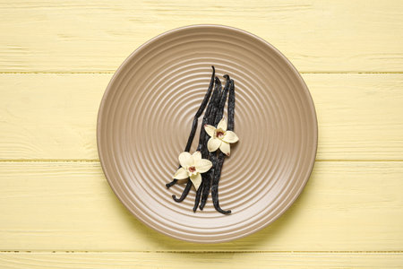 Plate with aromatic vanilla sticks and flowers on yellow wooden background