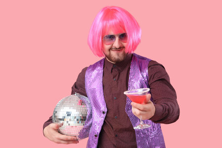 Handsome young man in wig with disco ball and glass of cocktail on pink backgroundの写真素材