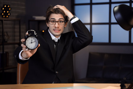 Young businessman with alarm clock trying to meet deadline in office at nightの写真素材