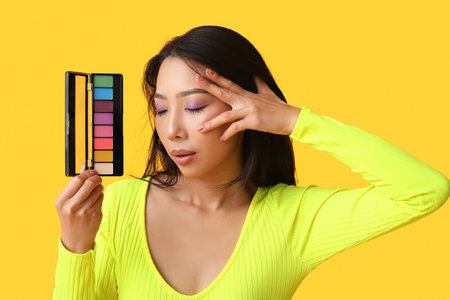 Young Asian woman with palette of eyeshadows on yellow backgroundの写真素材