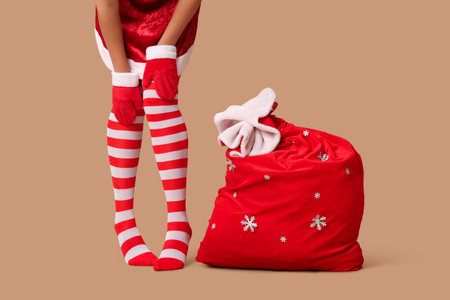 Woman dressed in striped stockings with bag of Christmas gifts on beige background