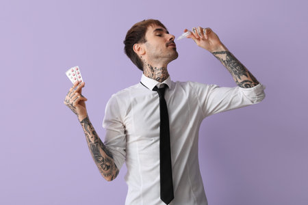 Allergic young man with pills using nasal drops on lilac background