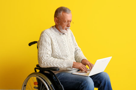 Senior man in wheelchair using laptop on yellow background