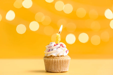 Tasty Birthday cupcake with burning candle on yellow table against blurred lightsの写真素材