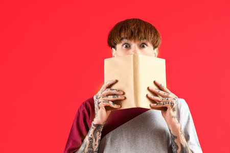 Afraid young tattooed man with book on red backgroundの写真素材