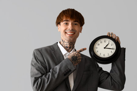 Handsome young tattooed businessman pointing at clock on gray background
