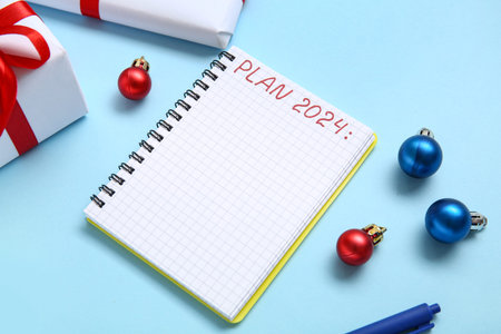 Notebook with text PLAN 2024, pen, Christmas balls and gift boxes on blue background, closeup