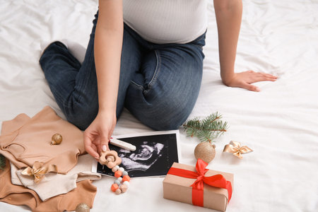 Pregnant woman with sonogram image, test, baby clothes and Christmas decorations on bed