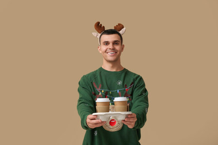 Young man in reindeer horns with paper cups of coffee on beige backgroundの写真素材