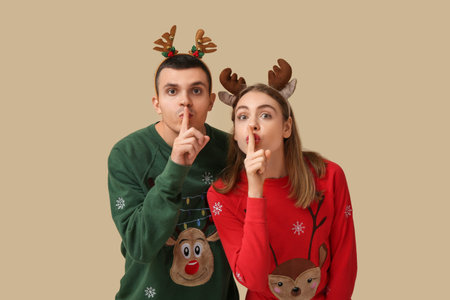 Beautiful young couple in reindeer horns showing silence gesture on beige background
