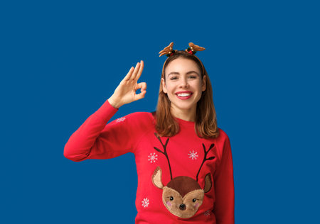 Beautiful young woman in reindeer horns showing ok gesture on blue backgroundの写真素材