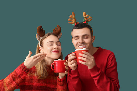 Beautiful young couple in reindeer horns holding cups of coffee with marshmallows on green backgroundの写真素材