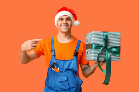 Male plumber in Santa hat with tools pointing at gift box on orange backgroundの写真素材